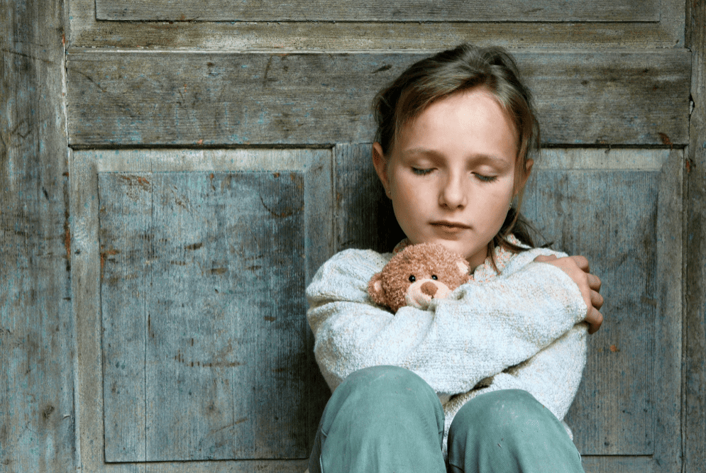 Niña de 8 años sentada en el suelo y recostada en una puerta mientras abraza su osito con los ojos cerrados